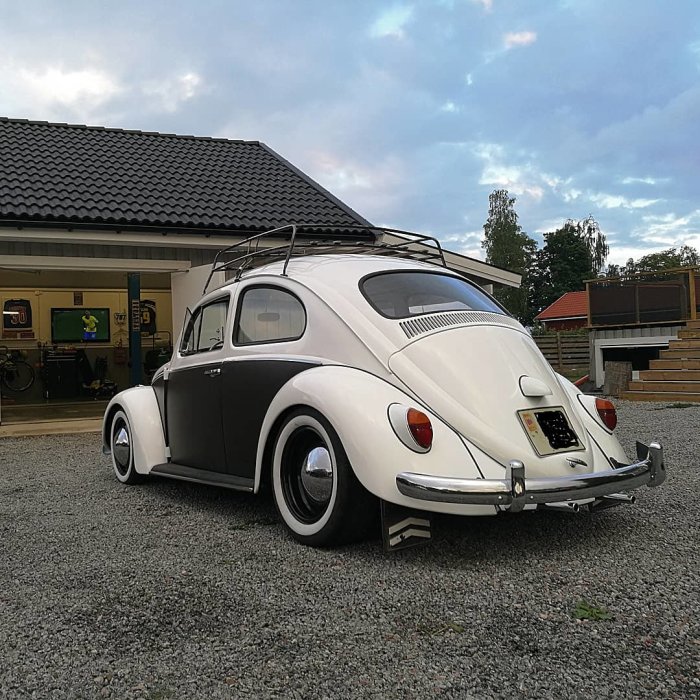 Vit och grå Volkswagen bubbla från 1962 med takräcke parkerad framför ett hus.