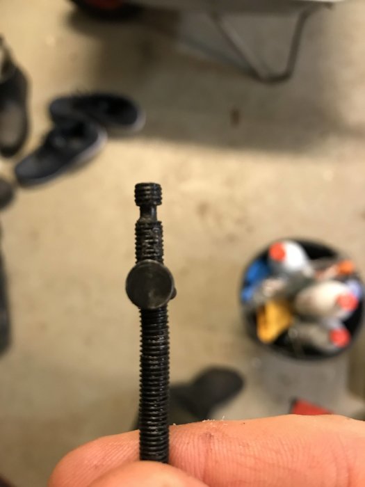 Böjd justeringsskruv för motorsågskedjespänning hållen framför ett suddigt garagegolv med skor i bakgrunden.