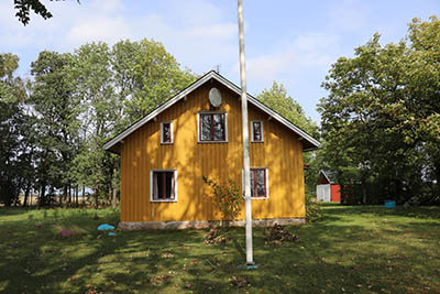 Gult trähus med enkelfönster och orenoverad fasad mot en bakgrund av träd och gräsmatta.