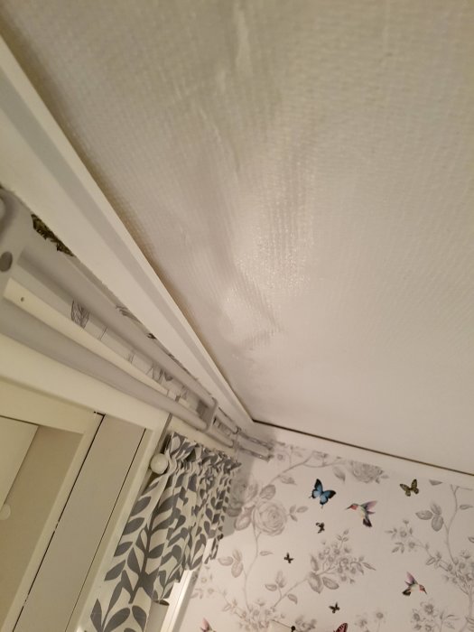 Sagging wallpaper in a ceiling corner with visible bulges and a peeled section exposing underlying surface.