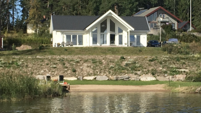 Husfasad med stora vikbara glaspartier och gavelfönster vid en strandkant.