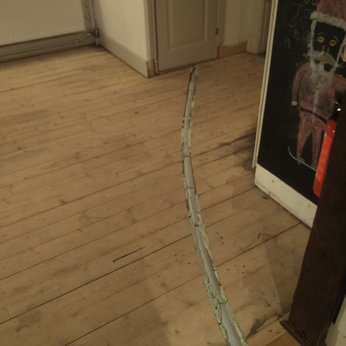Gammalt trägolv i kök med en fräst ränna som innehåller en isolerad vattenledning, behov av renovering.