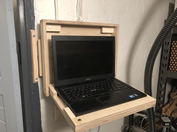 Hållare i trä för en bärbar dator monterad på en verkstadsvägg, med en Dell laptop öppen på hållaren.