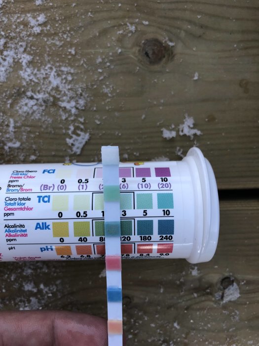 Teststicka som visar vattenkvalitetsvärden mot en flaska med färgkarta, placerad på ett snötäckt trägolv.