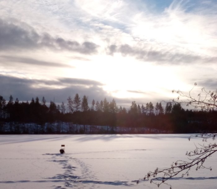 Solen bryter igenom molnen över en snötäckt sjö med träd i bakgrunden och spår i snön fram till en pulka.