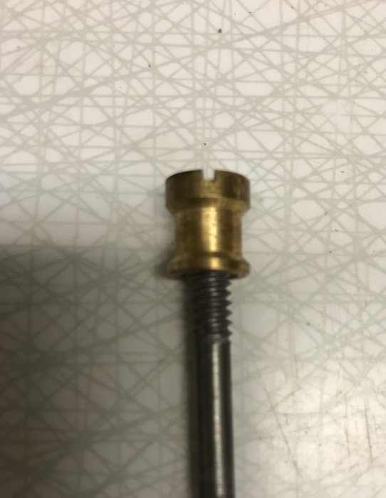 En skruv med en mässingsknopp och gängad axel mot en bakgrund med geometriskt mönster.