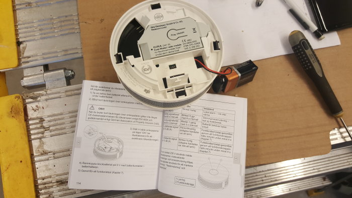 Rökdetektor med borttaget lock och batteri, instruktionsbok och verktyg på arbetsbänk.