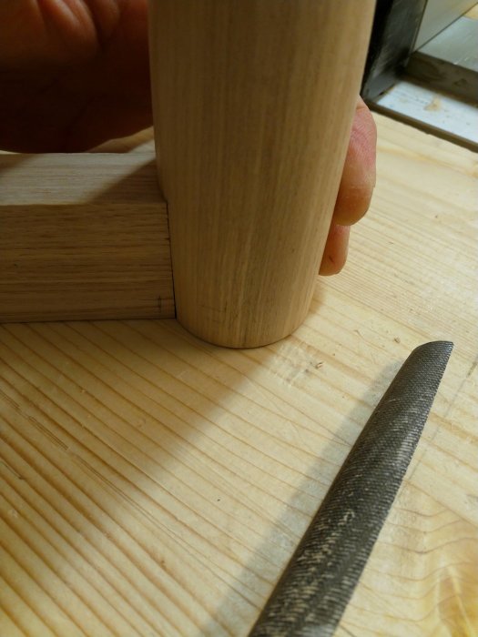 Hand som håller i ett hexagonalt utformat träben med markerade tapphål mot en träyta, intill ett stämjärn.