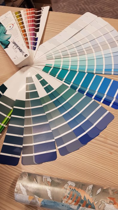 Färgkartor i blå och gröna nyanser bredvid en rulle tapet med mönster på bord.