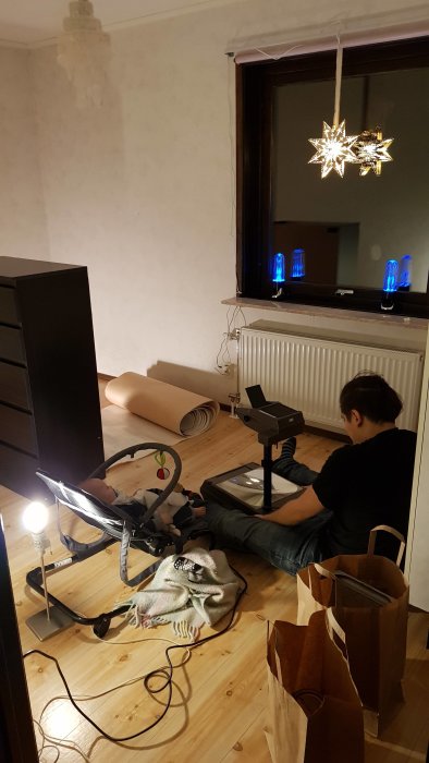 Person sitting on the floor preparing to hang wallpaper with tapet rolls, paint supplies, and a baby in a rocker nearby in a partially renovated room.