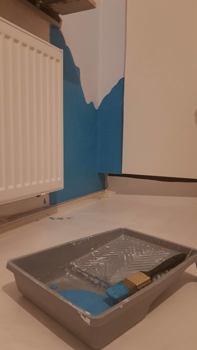 Målarprojekt i rum, väggparti halvmålat i blått med pensel och färgtråg i förgrunden.