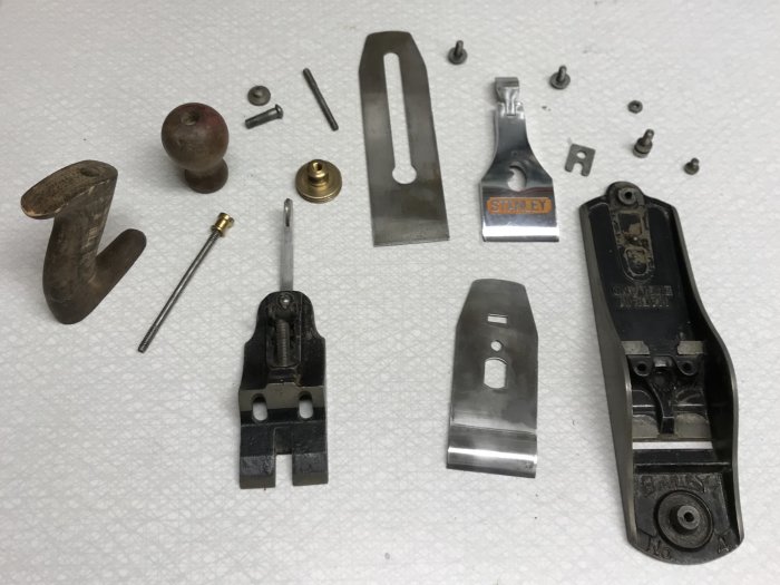 Delar av ett handverktyg, inklusive slipade metallkomponenter och ett trähandtag, spridda på en vit bakgrund.