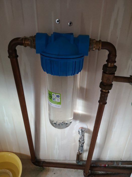 Installationsfärdigt vattenfilter i blå plastkoppling med genomskinlig behållare monterad på kopparledningar.