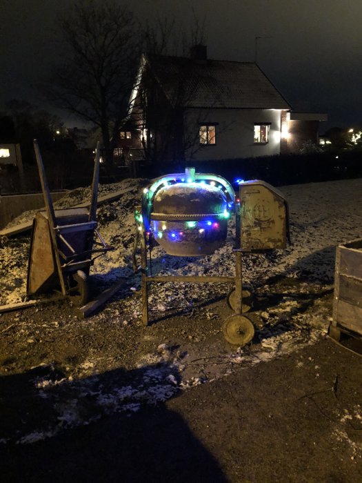 Julpynt bestående av färgglada ljusslingor upphängda på en gammal trädgårdsblandare i ett snöigt landskap på kvällen.