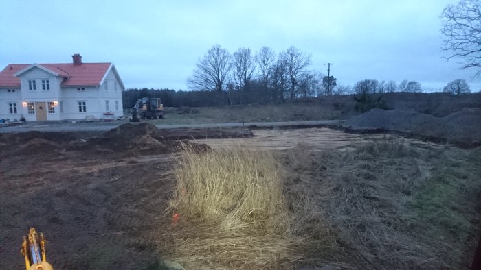 Schaktningsarbete vid uppförande av hus med grävmaskin, sandbäddar och grushögar i skymningen.