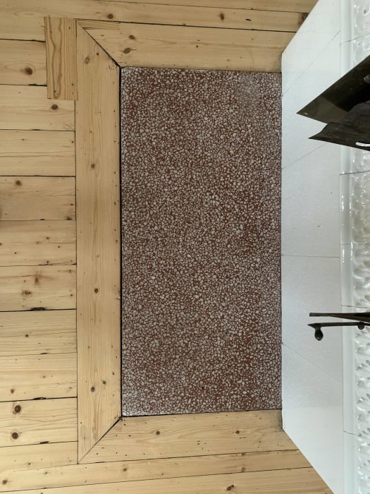 Rödaktig stenplatta framför vit kakelugn med trägolv och vit vägg i bakgrunden.