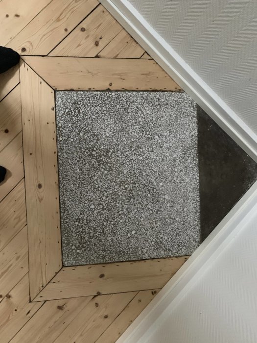Två stenplattor inbäddade i ett trägolv, en grå med små stenar och en mörk solid färg.