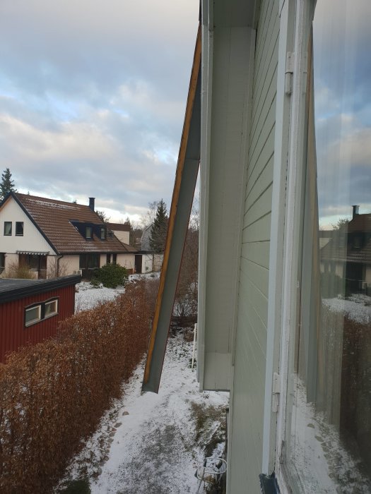 Skadad hushörna med en lossande gavelplatta, sett från fönstret med snöbelagd trädgård i bakgrunden.