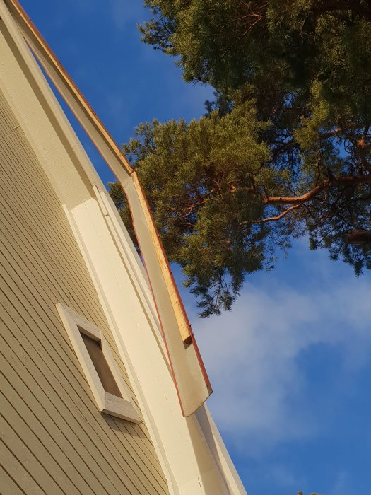 Vy från marken av ett hus hörn och tak mot en klarblå himmel med trädtoppar i bakgrunden.