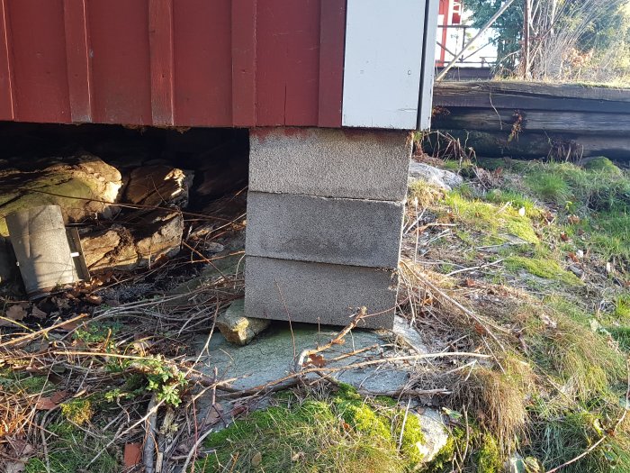 Osäker grund med betongblock staplade på brädbitar under en rödfärgad stuga med synligt bergrund.