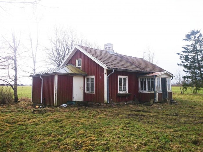 Röd trähus med veranda på en slätt, omgiven av några träd och öppen mark.