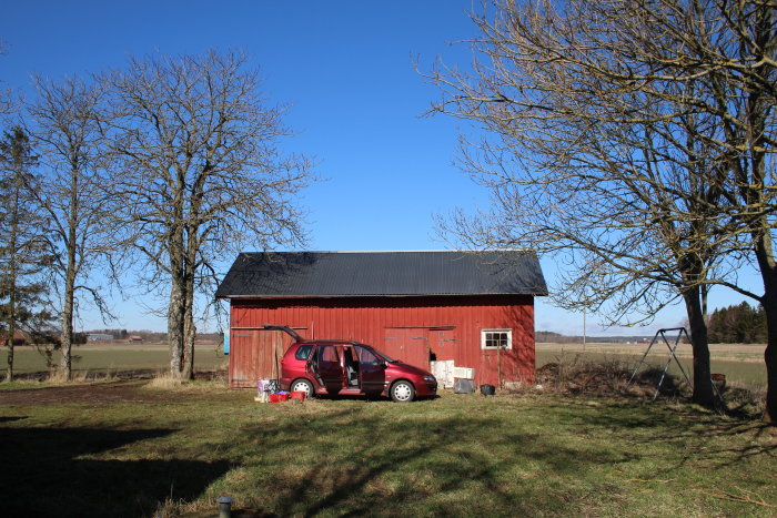Röd lada och röd bil framför, på en öppen åker vid två träd utan löv, blå himmel overhead.