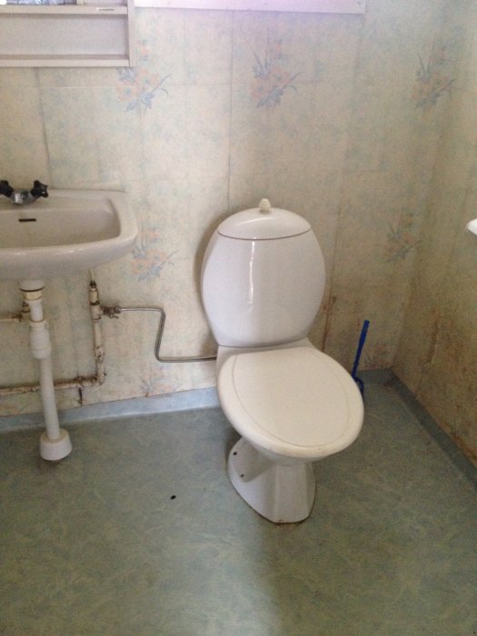Gammaldags toalett med blommig tapet och handfat i renoveringsbehov i ett äldre hus.
