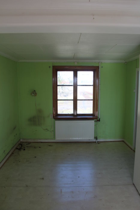 Ett grönt rum i behov av renovering med synliga vattenskador, ett fönster och en radiator.
