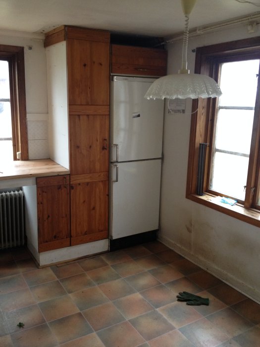 Gammalt kök med träskåp, vit kyl och fläckiga kakelplattor på golvet vid fönster och radiator.