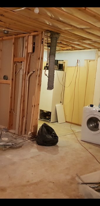 Spirorör i en orenoverad källarlokal som kommer genom takisoleringen, omgivet av byggmaterial och vitvaror.