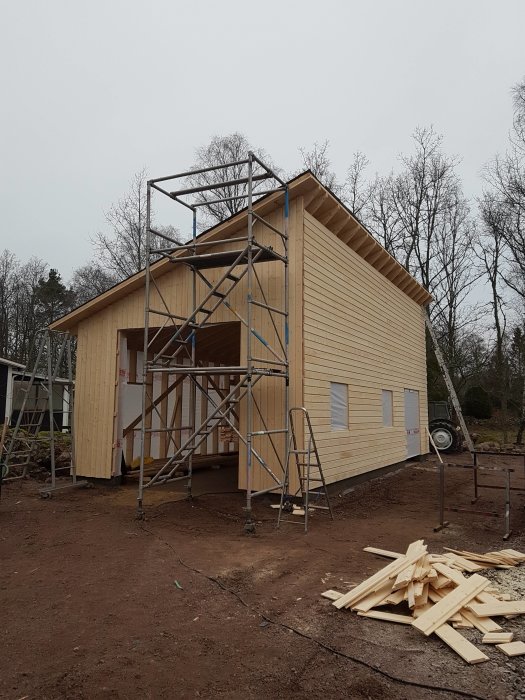 Ett garage under konstruktion med träskelett och fasad, byggställningar runtom, och träspill på marken.