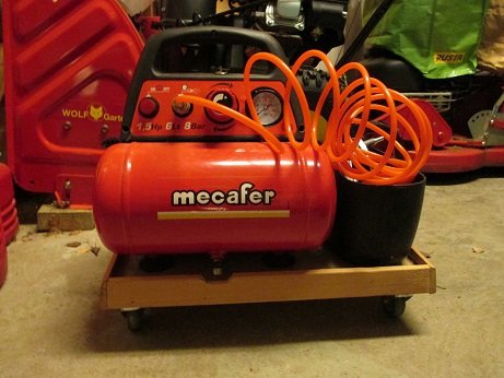 Liten röd kompressor på träplattform med länkhjul och orange luftslang, placerad i ett garage.