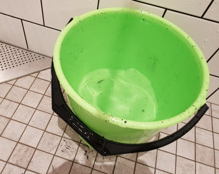 Grön hink fylld med vatten och svarta prickar mot en kaklad duschbakgrund.
