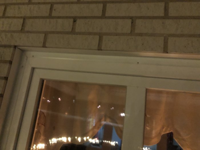 Nyinstallerade plåtar vid ett fönster under tegelvägg i ett hem med synlig gardin och utsikt mot exteriör belysning.