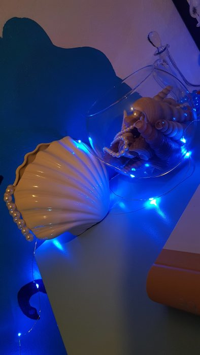 Mys-hörna med bläckfiskfigur och pärlor i en genomskinlig skål, bredvid en skalformad lampa, omgivet av blåa LED-ljus.