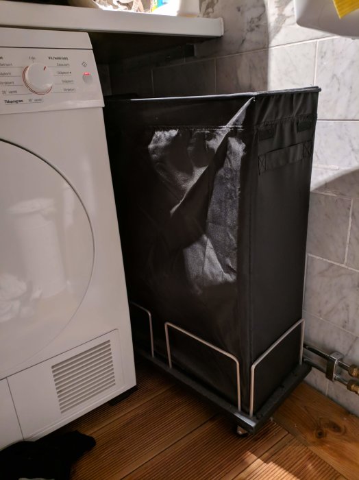 Svart tvättkorg i styvt tyg på hemmagjord vit rullvagn intill tvättmaskin och vitvaror.