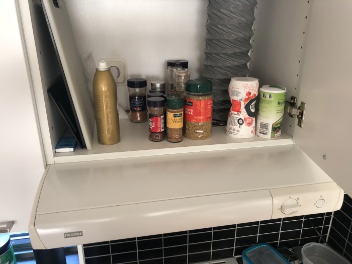 Gammal vit fläktkåpa under köksskåp med kryddburkar och teburkar på hyllan ovanför.