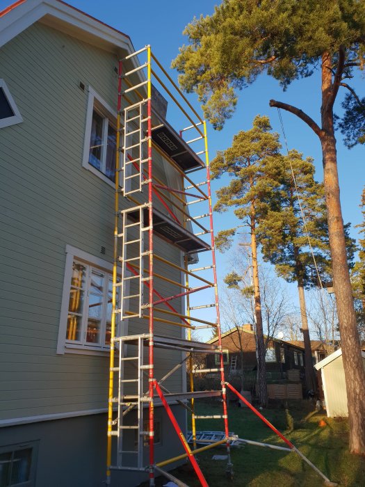 Ställning uppställd bredvid ett tvåvåningshus för renoveringsarbete, omgivet av träd och klar himmel.