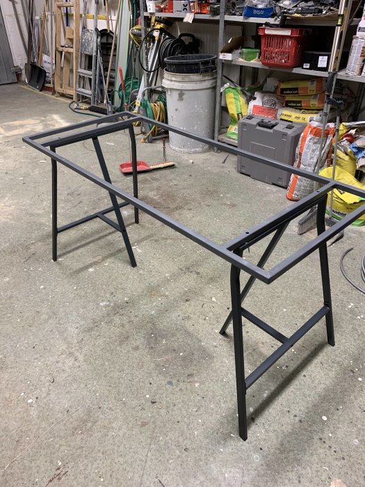Svetsad metallram för bord står i en verkstad med diverse verktyg och material i bakgrunden.