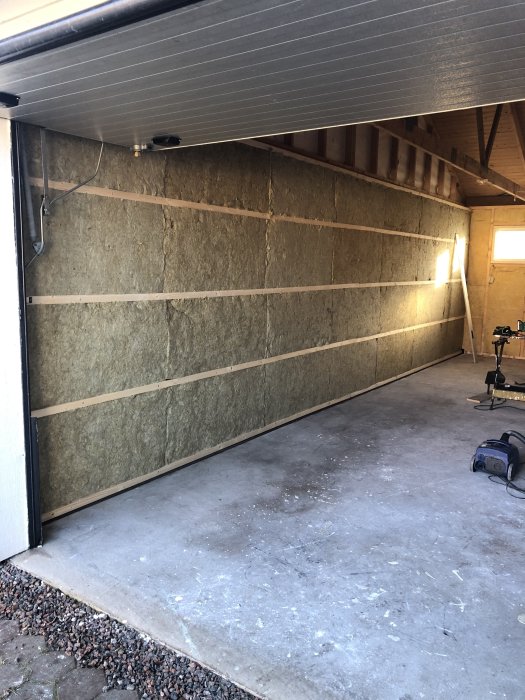 Garage under ombyggnad med synliga väggisoleringar och träbjälkar, oisolerat betonggolv och verktyg.