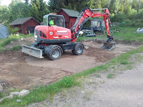 Röd Neuson grävmaskin vid grävarbete i en tomt med grävd jord och vegetation i bakgrunden.