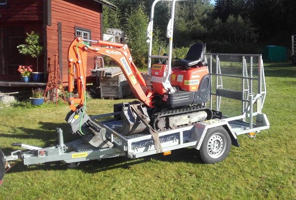 Liten orange Kubota-grävmaskin på en släpvagn framför en röd stuga i soligt gräsmiljö.