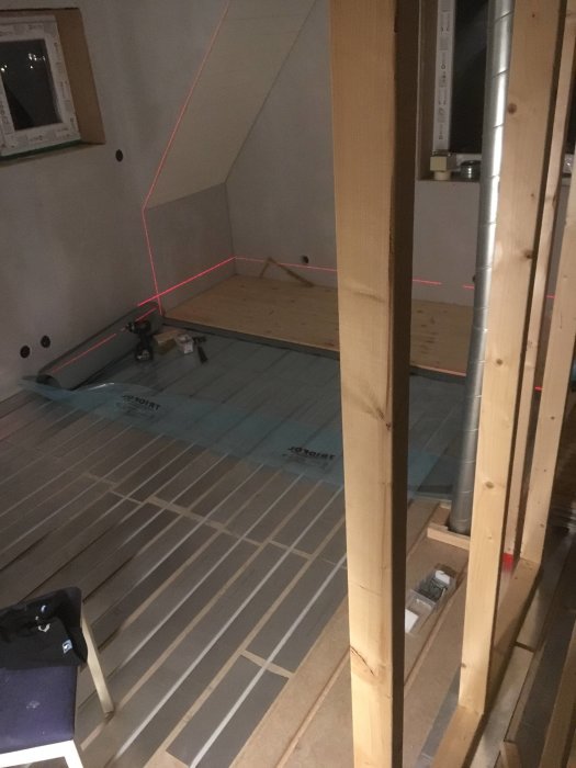 Installation av trägolv pågår i ett rum med mittvägg och synliga ventilationströr, laserlinje markerar väggen.