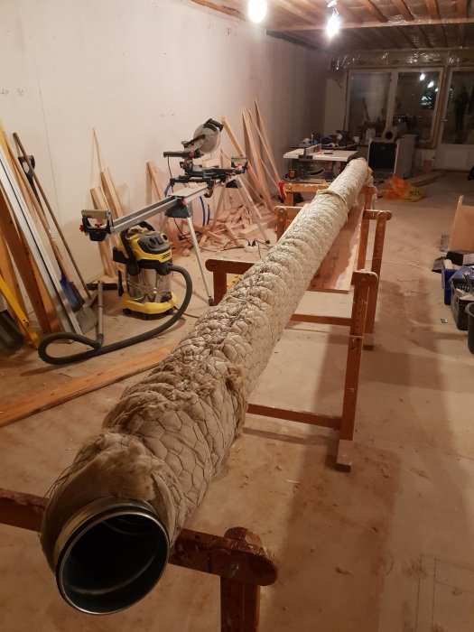 Isolerad ventilationsrör ligger på arbetsbänkar i en källarverkstad med virke och snickarverktyg i bakgrunden.