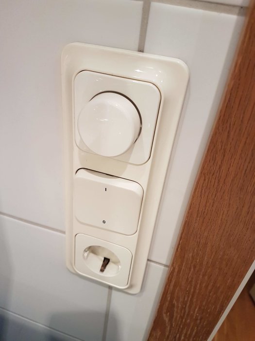 Vit väggmonterad vriddimmer med två knappar och ett eluttag i ett badrum.