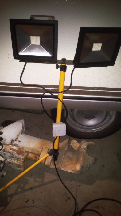 Två 50W arbetslampor monterade på en gul stång med kablar liggande på ett garagegolv.