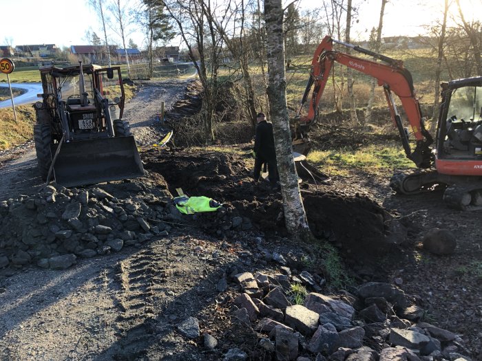 Grävmaskiner och en person arbetar med att dra fram el och fiber på en solig vinterdag, stenig mark synlig.