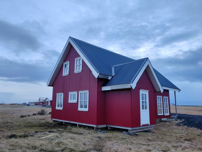 Rödmålat 1,5-planshus med vita fönster, dörrar och tak, beläget på en öppen isländsk landsbygd.