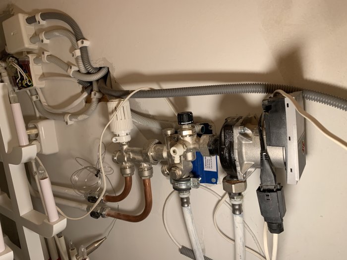 Äldre vattenburet värmesystem med synliga kopparrör, ventiler och en cirkulationspump installerade på en vägg.