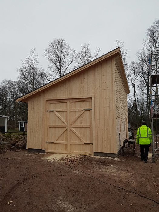 En nybyggd trägarage med stora dubbeldörrar under konstruktion, en person i reflexväst syns.